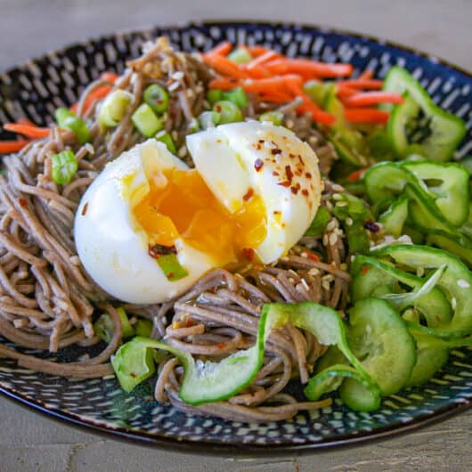 A soft boiled egg with runny yolk on top of a cold soba noodle salad with miso mirin dressing