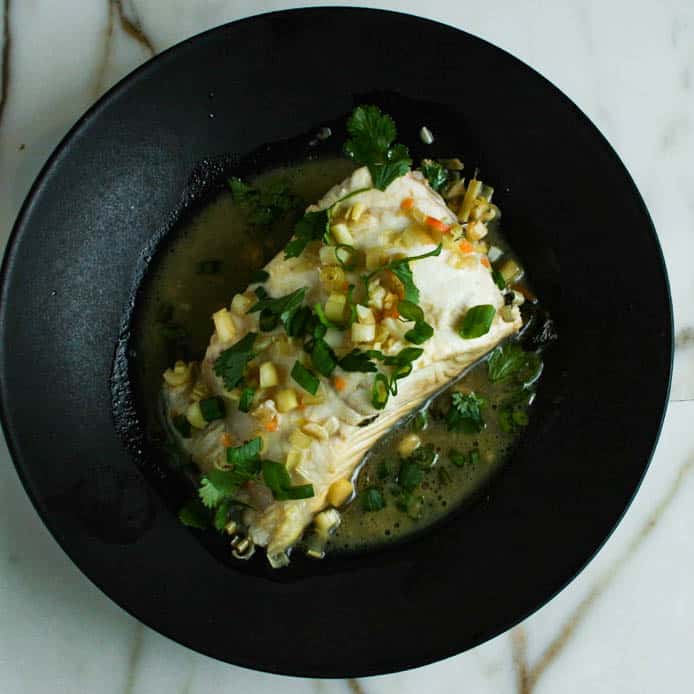 Overhead photograph of citrus and habanero poached sea bass. Photographed in a black bowl on top of marble. The sea bass is garnished with scallions and cilantro