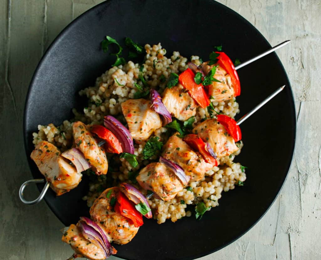 Greek Chicken Kebabs with red bell peppers and red onion served in a black bowl with couscous