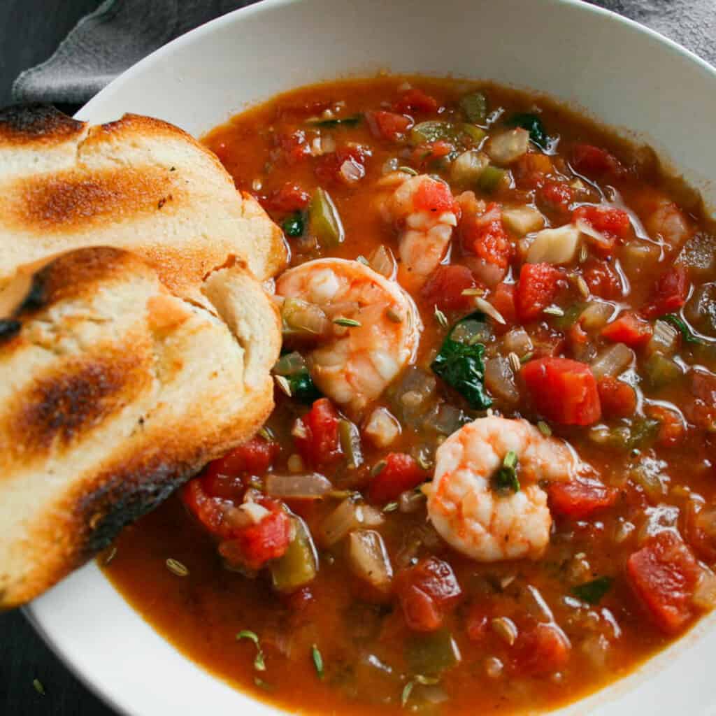 Seafood cioppino with toasted French bread. Photographed in a white bowl.