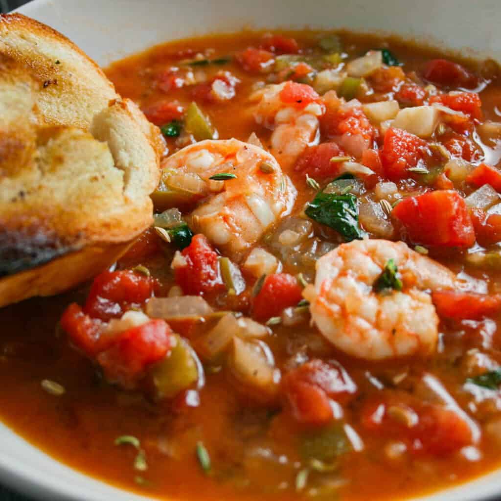 Seafood cioppino with toasted French bread. Photographed in a white bowl.