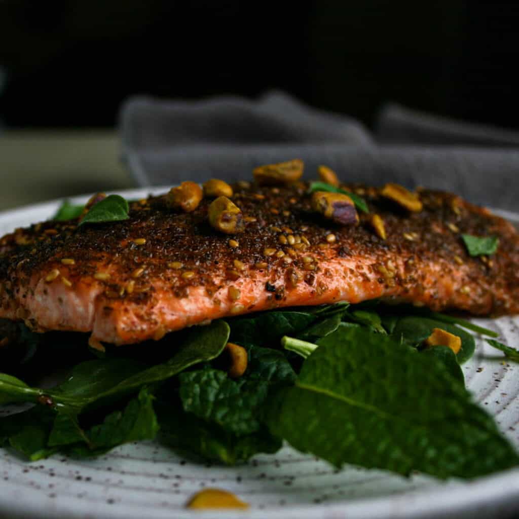 Eye Level photograph of za'atar crusted salmon on a bed of spinach and other greens. Garnished with fresh mint, oregano, and pistachios
