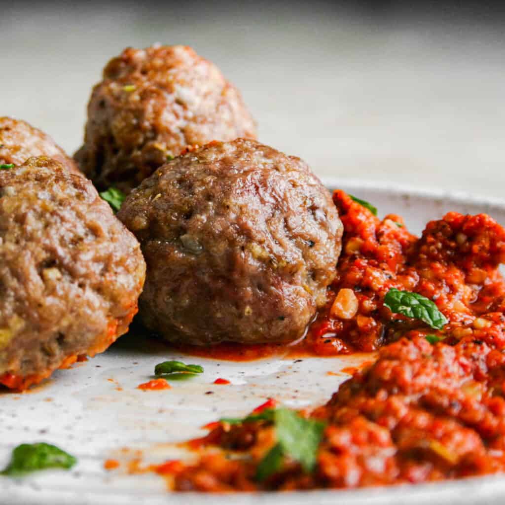 Lamb Meatballs served with Romesco Sauce. Photographed eye level on a white ceramic plate. Garnished with Mint Leaves