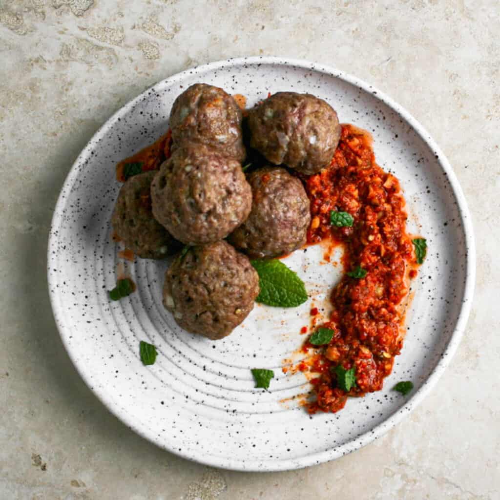 Overhead photograph of lamb meatballs with middle eastern inspired romesco. Photographed on a white ceramic plate on a travertine backdrop. Garnished with fresh mint leaves