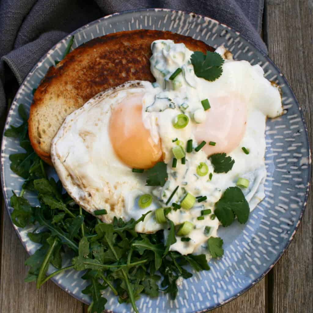 Fried Eggs with Miso Creme Fraiche. Finished with scallions and chives. served over sourdough bread and a side salad. Feast Local. Photographed on a blue and white plate.