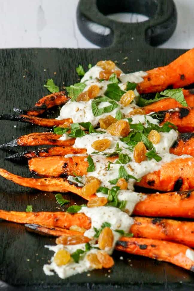 Grilled Mediterranean Carrots with a creamy yogurt sauce. Toped with fresh mint and golden raisins. Served on a black wood cutting board. 