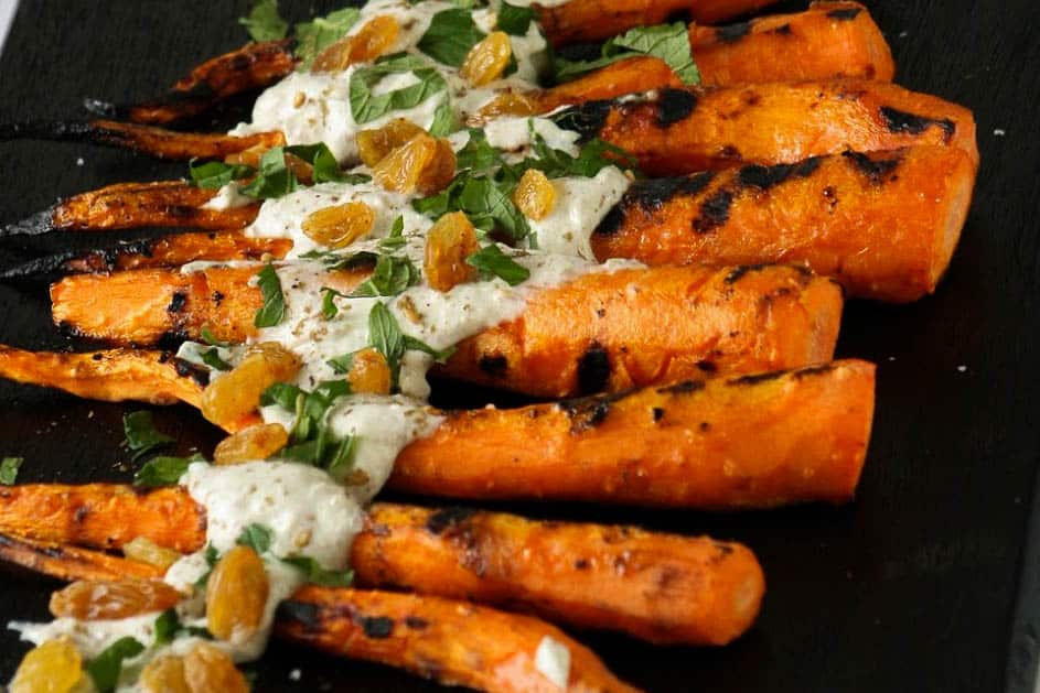 Hero Shot of Grilled Mediterranean Carrot. Shows grilled carrots with a yogurt sauce, fresh mint, and golden raisins. Photographed on a black cutting board. 