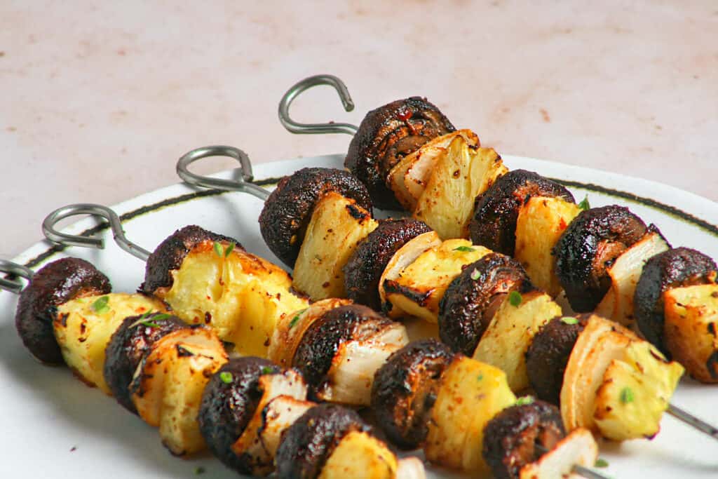Hero Photograph of Vegan Jamaican Jerk Mushroom Skewers. Photographed on the skewers on a white plate on a stone table