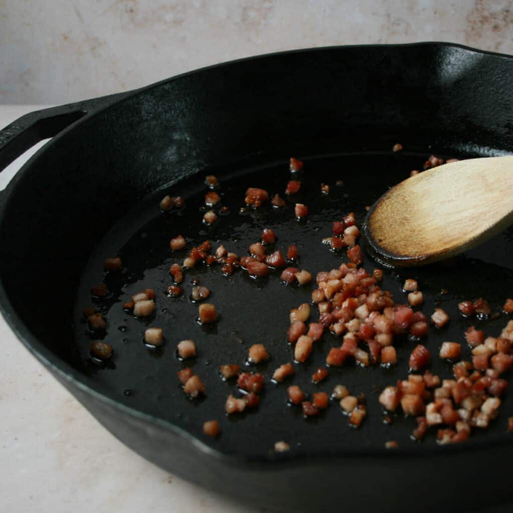 Photograph of cubed pancetta being cooking in a cast iron skillet. A wooden spoon is used to stir. 
