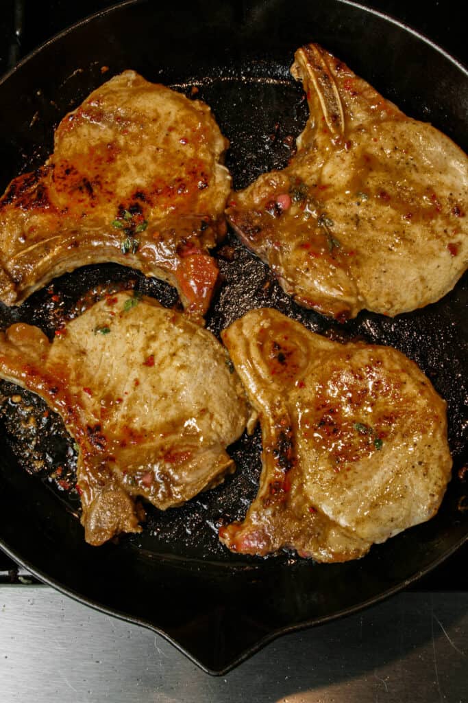 Pan Seared Maple Pork Chop photographed cooking in a cast iron Staub skillet. the skillet is black. there is a maple sauce on the pork chops