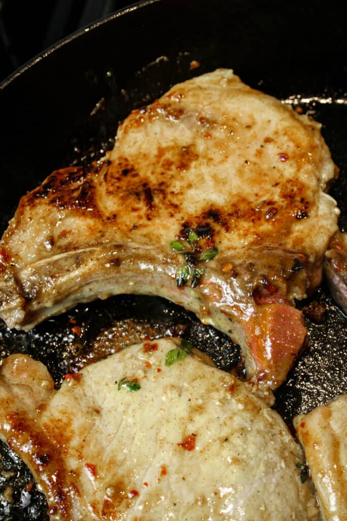 Pan Seared Maple Pork Chop photographed cooking in a cast iron Staub skillet. the skillet is black. there is a maple sauce on the pork chops