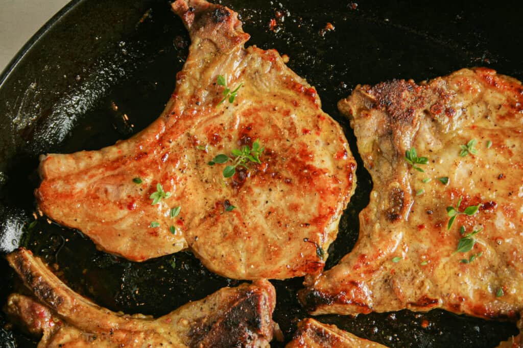 Pan Seared Maple Pork Chop photographed cooking in a cast iron Staub skillet. the skillet is black. there is a maple sauce on the pork chops