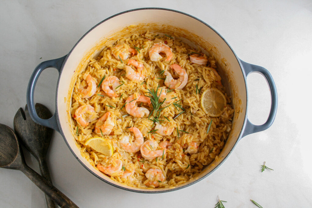 Hero photograph of creamy rosemary orzo with shrimp. Photographed in a steel blue dutch oven with wooden serving utensils on the side.
